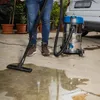 A person is using a wet/dry vacuum cleaner to clean up a spill on a concrete floor, with plants and a car in the background. Produktbild-Vorschau