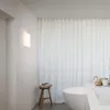 A modern bathroom with a bathtub, a stool with books, and a wall lamp. Produktbild-Vorschau