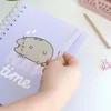 A hand is holding a purple notebook with a cartoon cat on it, with a pencil and binder clips in the background. Náhľad obrázku produktu