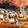 Several meerkat keychains are displayed on a sandy surface with a blurred desert-like background. Produktbild-Vorschau