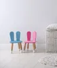 Two children's chairs with bunny ears, one blue and one pink, are placed on a light wooden floor in front of a white wall. A woven basket with a striped cushion is to the right. Produktbild-Vorschau
