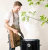 A man wearing a brown leather apron is grilling outdoors, with smoke rising from the grill. Produktbild-Vorschau