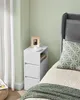 A white bedside table with drawers next to a bed with a grey headboard and a patterned pillow. Náhled obrázku produktu