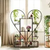 A heart-shaped plant stand filled with various plants and books, placed near a window with curtains. Produktbild-Vorschau