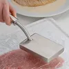 A hand is using a meat tenderizer to flatten a piece of meat wrapped in plastic. In the background, a plate with breaded meat is visible. Podgląd zdjęcia produktu