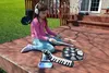A young girl wearing headphones is playing a keyboard and drum set on a wooden deck outdoors. Produktbild-Vorschau