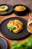 A close-up of a plate of pasta with basil and a glass of white wine, with other dishes in the background. Náhľad obrázku produktu