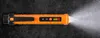 A close-up, top-down view of an orange and black non-contact voltage tester with its light on, lying on a dark, textured surface. Podgląd zdjęcia produktu