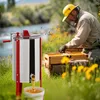 A beekeeper in protective gear works with a beehive next to a honey extractor, with a river and foliage in the background. Produktbild-Vorschau