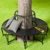 A black metal garden bench is placed around a tree trunk on a green lawn. Produktbild-Vorschau