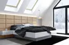 A modern bedroom with a bed against a padded headboard, under slanted ceilings with skylights, and a large window showing a winter landscape. Podgląd zdjęcia produktu