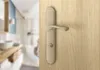 A close-up of a door handle on a wooden door, with a blurred bathroom in the background. Produktbild-Vorschau