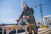 A construction worker is using a circular saw on a rooftop with a crane in the background. Podgląd zdjęcia produktu