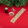A collection of Harry Potter-themed stationery items, including a ruler, a stamp, glasses charm, and a golden snitch-like object, arranged on a red surface with a hint of Christmas tree branches in the background. Náhled obrázku produktu