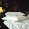 Two white pillows are stacked on top of each other, with a duvet partially visible underneath. A lamp is on the left, and a dark floral wallpaper is in the background. Produktbild-Vorschau