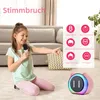 A young girl is sitting on the floor, holding a microphone and pointing, with a pink karaoke machine in the foreground. The background shows a living room setting. Produktbild-Vorschau