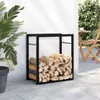 A black metal firewood rack filled with logs, placed on a patio next to a white wall and a large potted plant. A wooden chair and table are visible in the background. Náhled obrázku produktu