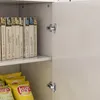 A close-up view of an open white cabinet, showing books on a shelf and bags of chips below. The cabinet doors have silver hinges. Podgląd zdjęcia produktu