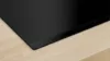A close-up view of the corner of a black induction hob resting on a light wood surface. Produktbild-Vorschau