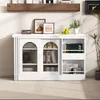 A white cabinet with sliding glass doors, filled with books and decorative items, is placed on a carpeted floor against a white wall. A wooden shelf with books is mounted above it. Produktbild-Vorschau