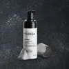 A bottle of Filorga Skin-Prep Enzymatic Cleansing Foam with some foam dispensed next to it, on a dark, textured surface. Podgląd zdjęcia produktu