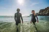 Two people in wetsuits walking into the ocean on a sunny day. Náhľad obrázku produktu
