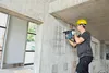 A construction worker is using a Bosch rotary hammer drill to drill into a concrete wall. The worker is wearing a yellow hard hat and safety glasses. The wall has a grid of lines drawn on it, indicating where to drill. There is an open doorway to the left, Produktbild-Vorschau