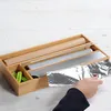 A hand is pulling aluminum foil from a bamboo dispenser that holds rolls of parchment paper, aluminum foil, and plastic wrap. Produktbild-Vorschau