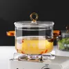 A glass pot with a golden handle and amber handles is placed on a small portable stove with a blue flame. Produktbild-Vorschau