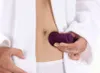 A person in a white robe is holding a purple massage tool to their abdomen. Produktbild-Vorschau