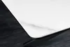 Close-up of the corner of a white ceramic table with a marble pattern and a black edge, resting on a dark, textured surface. Produktbild-Vorschau