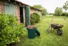 A green trash bin filled with garden debris sits on a grassy lawn next to a wheelbarrow and shovel, with a brick building in the background. Podgląd zdjęcia produktu