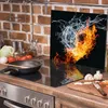 A kitchen counter with a stovetop, a cutting board with vegetables, and a glass splashback with a fire and water design. Produktbild-Vorschau