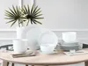 A white porcelain dinnerware set is arranged on a light wooden table, with a vase of green decorative leaves in the background. Produktbild-Vorschau
