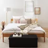 A bedroom scene with a bed, pillows, blankets, a floor lamp, and a black storage bench in front of the bed. Produktbild-Vorschau