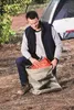 A man is kneeling on the ground, packing an orange item into a tan bag. A tent is visible in the background. Podgląd zdjęcia produktu