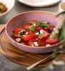 A close-up of a tomato and feta salad in a pink bowl, served on a wooden tray with a fork. Náhled obrázku produktu