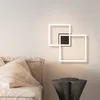 A modern wall lamp with two overlapping square frames is mounted on a light-colored wall above a sofa and a side table. Voir les images du produit