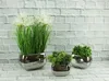 Three artificial plants in silver ceramic pots are displayed against a white brick wall. Podgląd zdjęcia produktu
