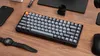 A mechanical keyboard is placed on a wooden desk alongside a smartphone, a watch, and a mouse. Produktbild-Vorschau