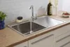 A stainless steel kitchen sink with a faucet and a draining board, set into a wooden countertop with white cabinets below. The background features white subway tiles. Produktbild-Vorschau
