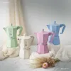 Four Bialetti Moka coffee makers in pastel colors (cream, green, pink, and blue) are arranged on a surface with draped fabric and dried flowers. Anteprima dell’immagine prodotto