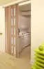 A folding door is partially open, revealing a small kitchen area with white cabinets and appliances. A bright green shelf with an apple is in the foreground. Podgląd zdjęcia produktu