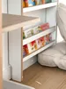 A close-up view of a white wooden children's bookshelf filled with colorful books, placed on a wooden floor next to a large floor cushion. Podgląd zdjęcia produktu