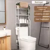A white bathroom shelf unit with multiple shelves is placed over a toilet. Various toiletries and a rolled towel are on the shelves. A brown towel is draped over the top shelf. Produktbild-Vorschau