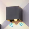 A modern, dark gray cube-shaped outdoor wall light is mounted on a light gray wall. Light emanates from the top and bottom of the fixture, casting shadows on the wall. Blue wavy lines indicate motion detection. Produktbild-Vorschau