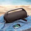 A black portable speaker is placed on a denim shirt on a sandy beach with the ocean in the background. A smartphone is charging from the speaker. Náhľad obrázku produktu
