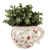 A ceramic planter shaped like a teacup, decorated with strawberries and vines, filled with green artificial plants. Podgląd zdjęcia produktu