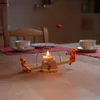 A candle holder with two figures on a seesaw, placed on a wooden table with cups and saucers in the background. Anteprima dell’immagine prodotto