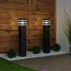 Two black bollard lights are positioned in a garden setting, with a wooden fence in the background and decorative white stones and green bushes around their bases. Produktbild-Vorschau